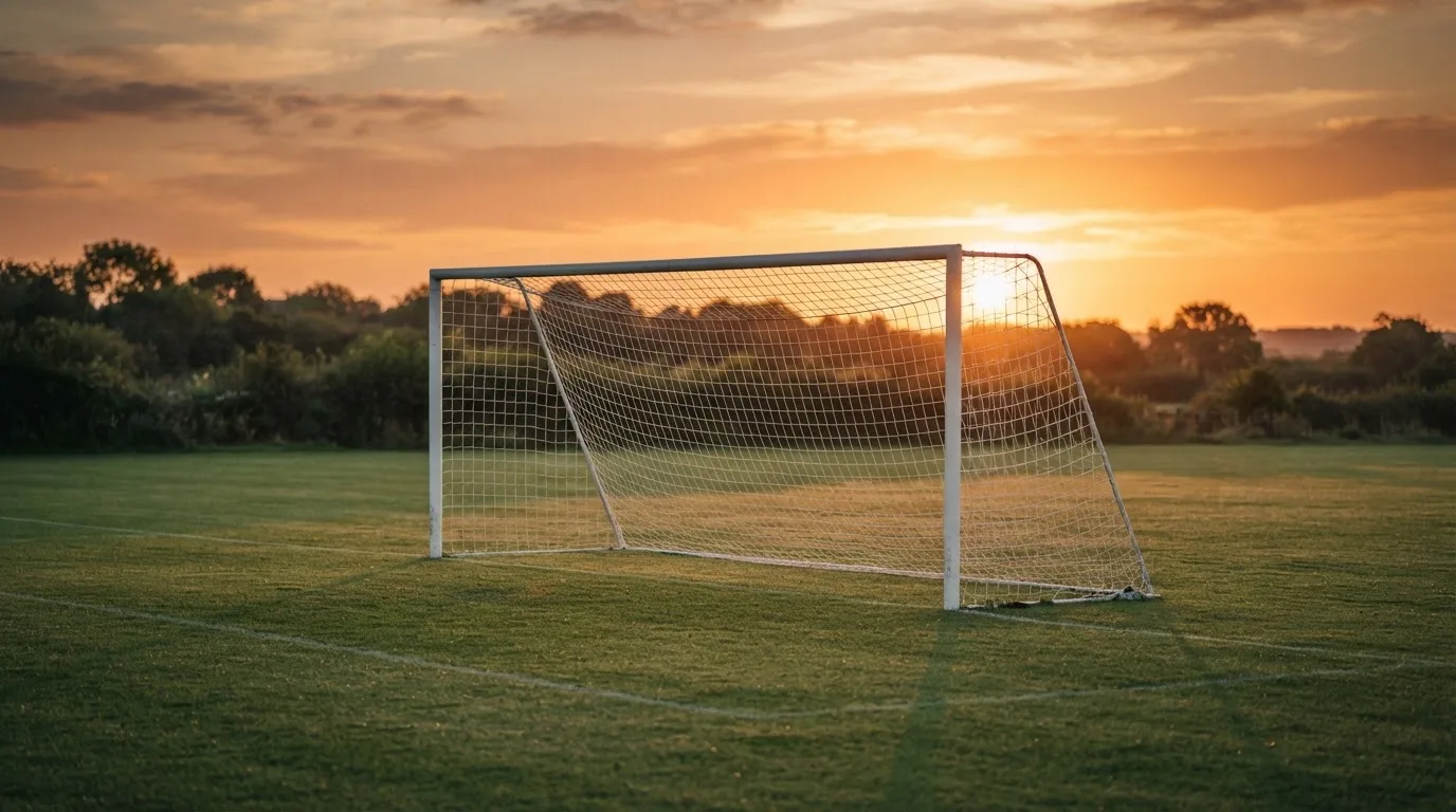 Ein Fußballtor mit Netz bei Sonnenuntergang auf einem leeren Platz, die Schatten der Torpfosten liegen lang auf dem Gras.
