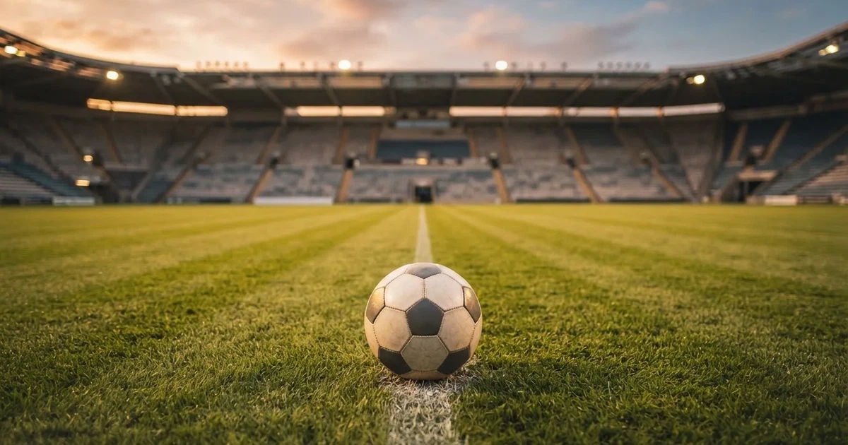 Ein Fußball liegt auf dem Anstoßpunkt eines leeren Rasenplatzes, im Hintergrund eine große Stadiontribüne im Abendlicht.