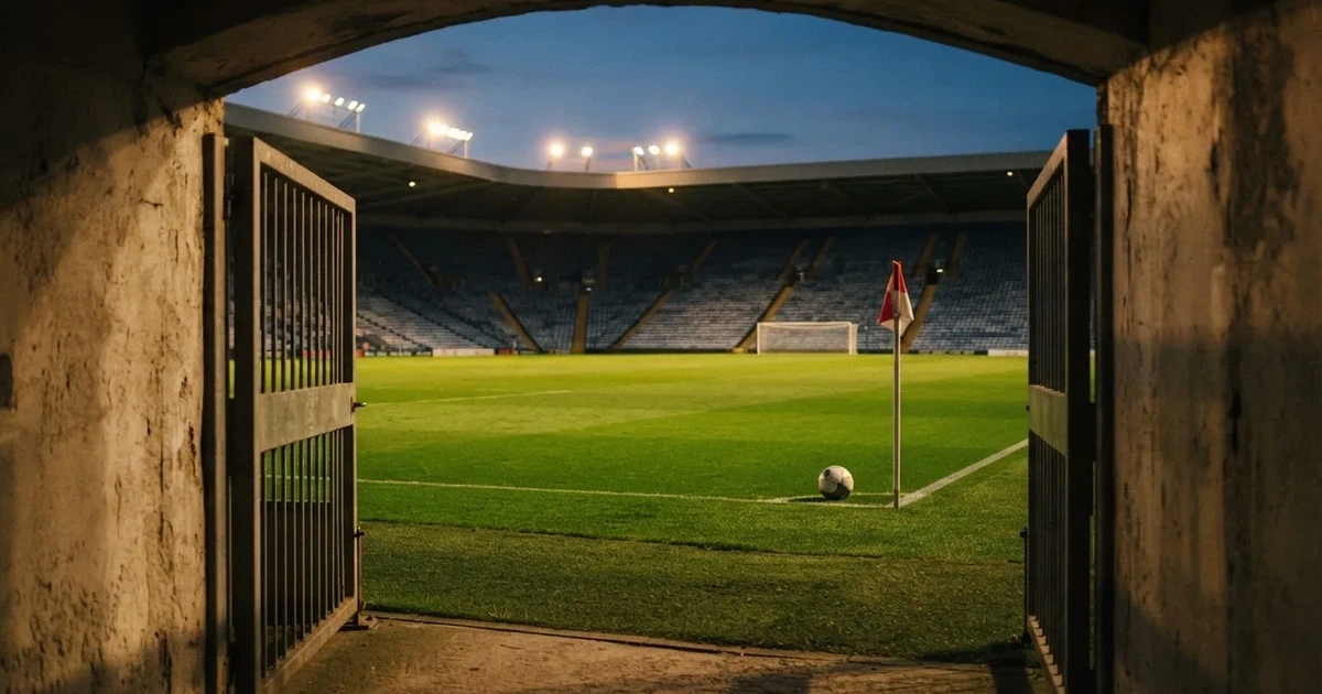 Blick durch ein Stadiontor auf einen grünen Rasen im Abendlicht, ein Ball ruht an der Eckfahne, die Tribünen sind leer.