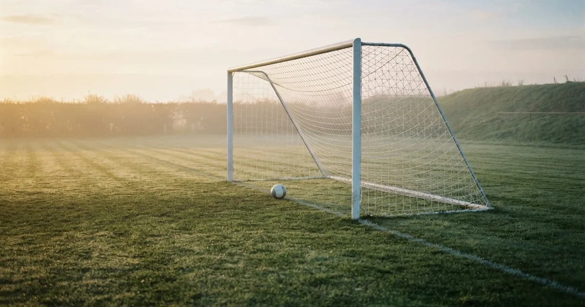 Ein Fußballtor mit gespanntem Netz steht auf einem leeren Rasenplatz, davor ein einzelner Ball, Morgendunst liegt über dem Feld.