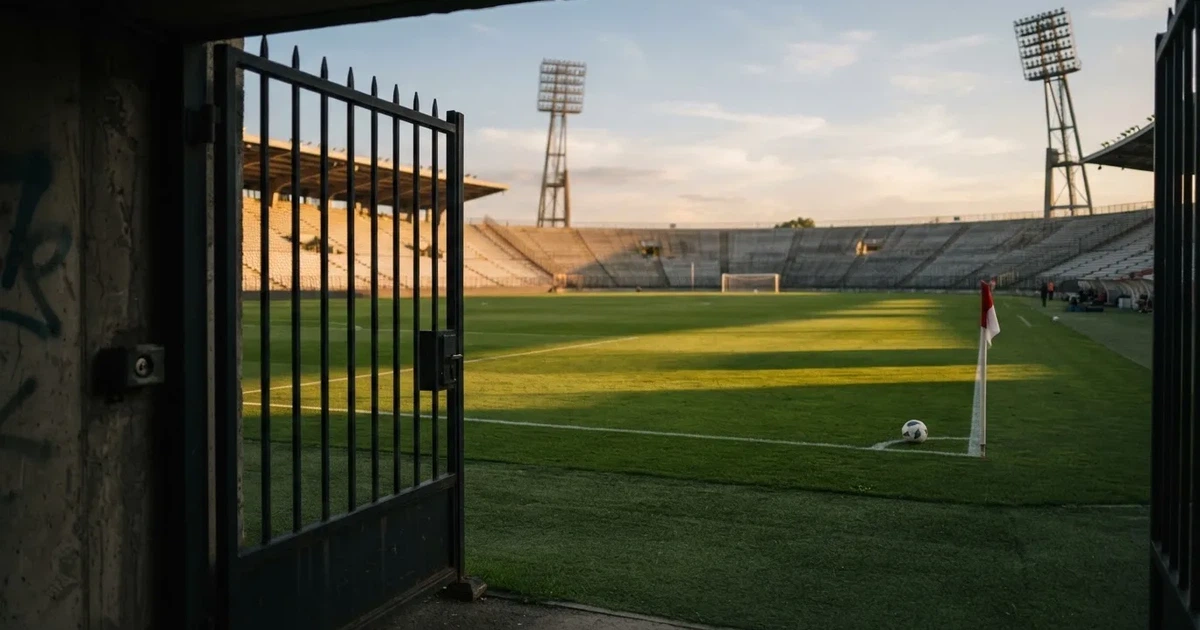 Blick durch ein Stadiontor auf einen grünen Rasen im Abendlicht, ein Ball liegt an der Eckfahne, die Ränge sind leer.
