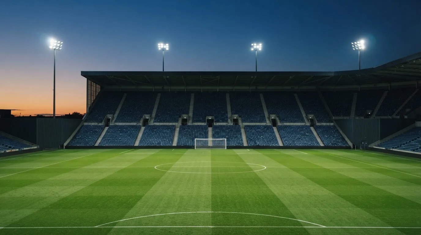 Eine leere Stadiontribüne unter Flutlicht aus der Ferne betrachtet, die Rasenfläche leuchtet grün in der Dämmerung.