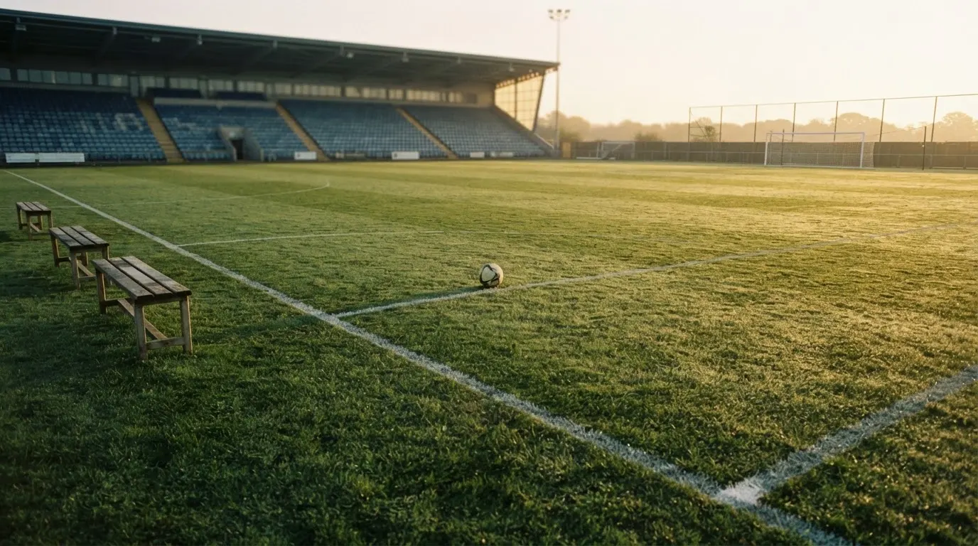 Ein Fußballplatz mit frisch gezogenen Kreidelinien im ersten Morgenlicht, leere Ersatzbänke am Spielfeldrand.