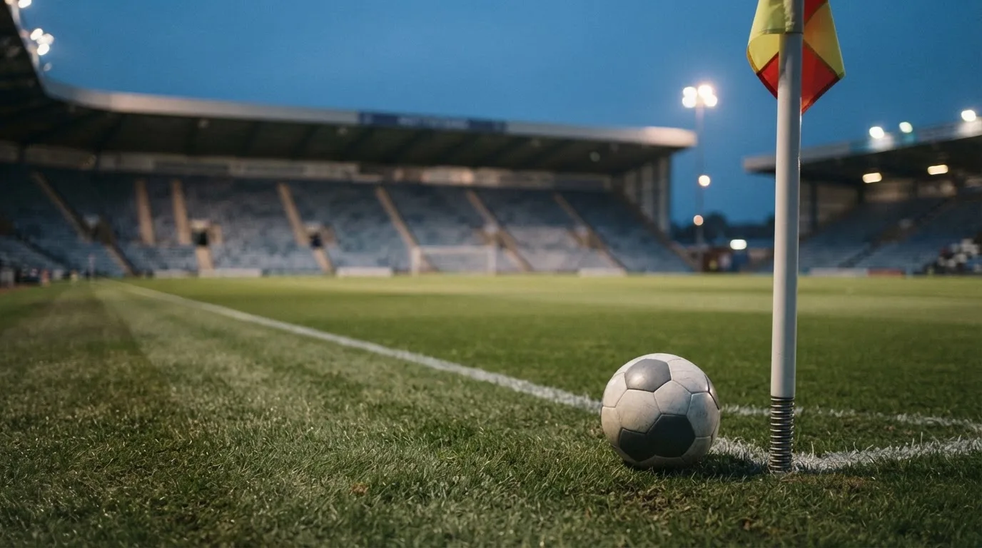 Ein Fußball liegt neben einem Eckfahnen-Pfosten auf einem Rasenplatz, im Hintergrund ein leeres Stadion in der Dämmerung.