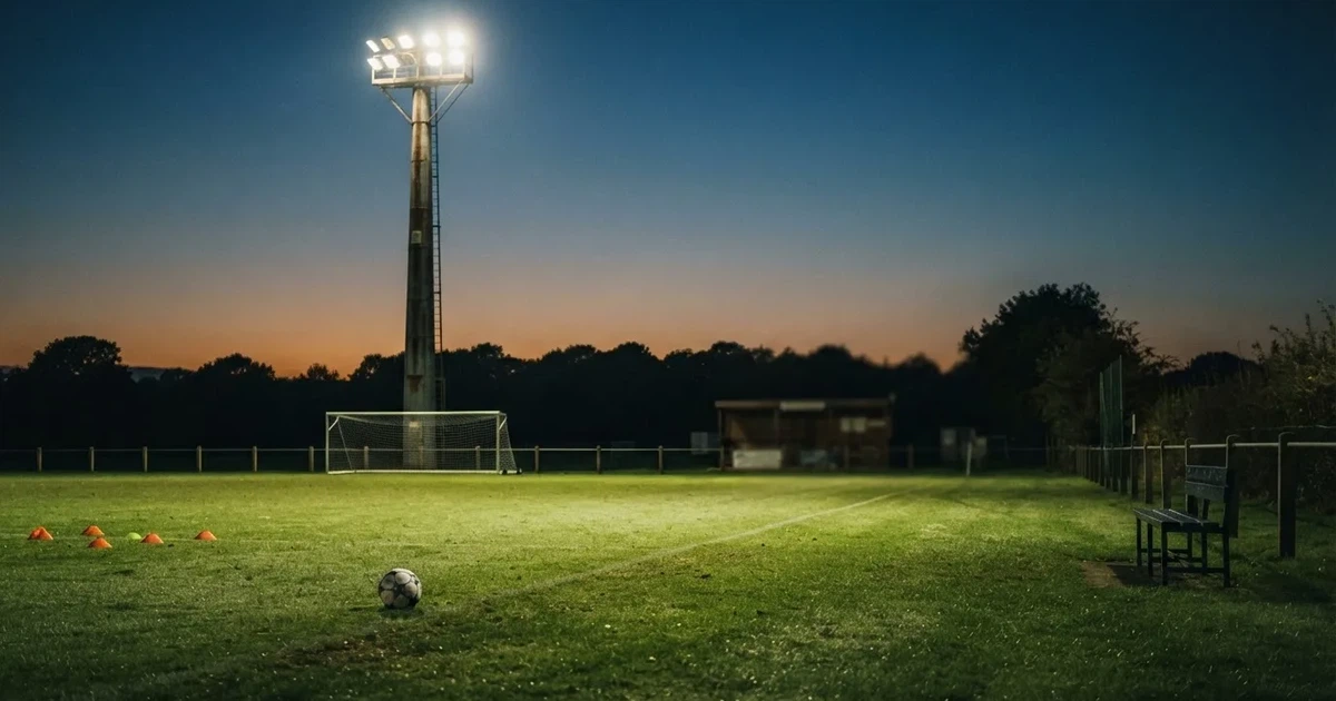 Eine Flutlichtanlage ragt über einen Trainingsplatz, auf dem Rasen liegen verstreut Hütchen und ein Ball, am Himmel die Abenddämmerung.