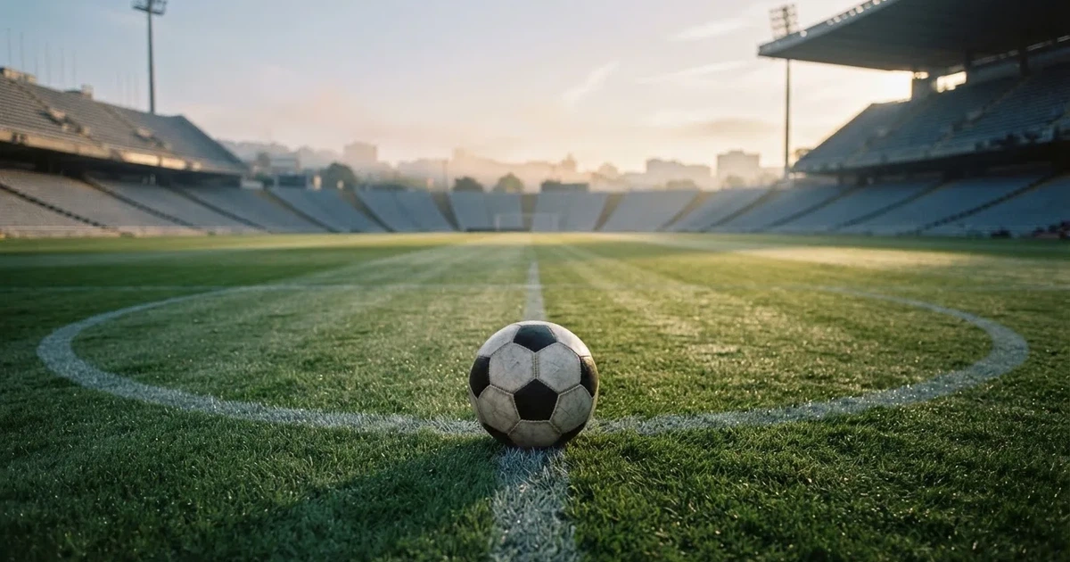 Ein Fußball liegt auf dem Mittelkreis eines Stadionrasens am frühen Morgen, im Hintergrund leere Ränge ohne erkennbare Beschriftung.