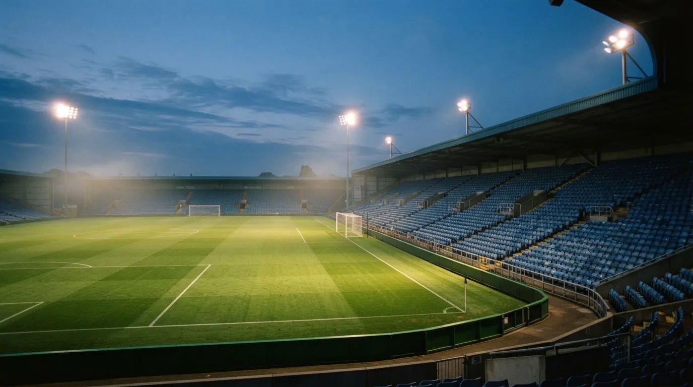 Eine leere Stadiontribüne unter Flutlicht aus der Ferne betrachtet, die Rasenfläche leuchtet grün in der Dämmerung.
