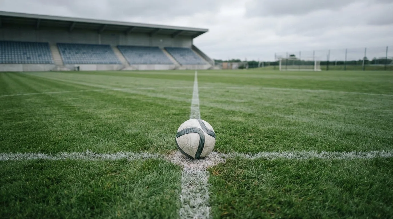 Ein Fußball liegt auf dem Anstoßpunkt eines leeren Rasenplatzes, im Hintergrund eine ruhige Tribüne unter bedecktem Himmel.