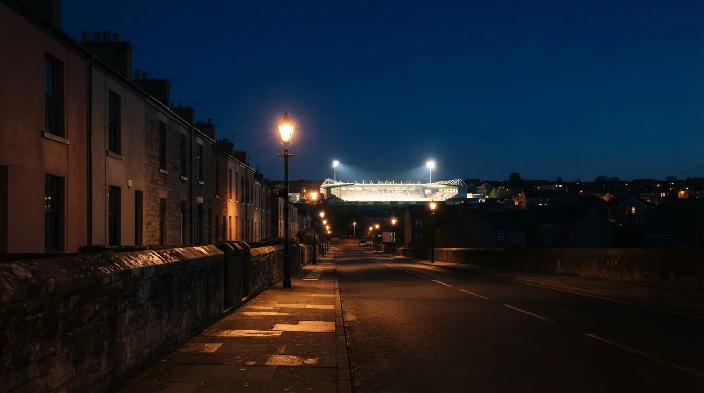 Ein Stadion unter Flutlicht aus der Ferne gesehen, davor eine dunkle Straße mit Laternen und ohne Menschen.