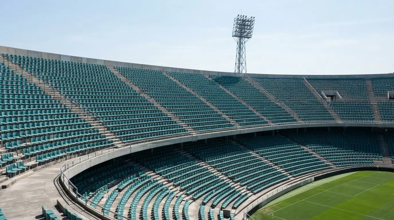 Eine leere Tribüne eines Fußballstadions bei Tageslicht, Sitzschalen in Reihen und dahinter ein Flutlichtmast.