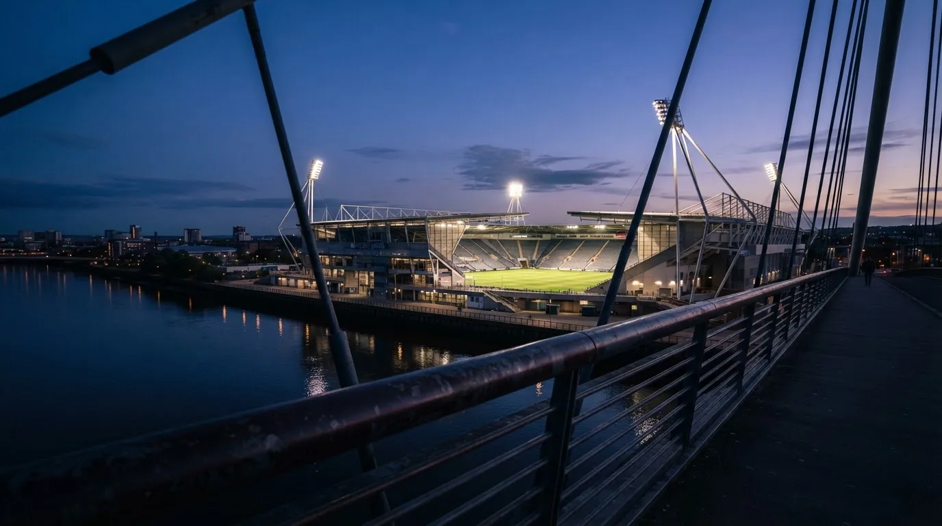 Ein Stadion bei Dämmerung mit eingeschalteten Flutlichtmasten, von einer Brücke aus aufgenommen.