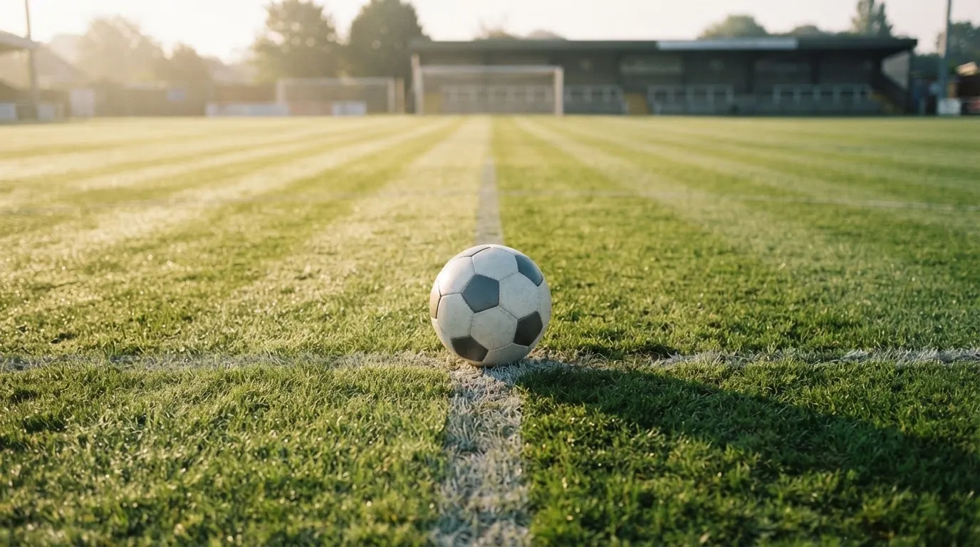 Ein Fußball liegt auf dem Anstoßkreis eines leeren Rasenplatzes im frühen Morgenlicht.