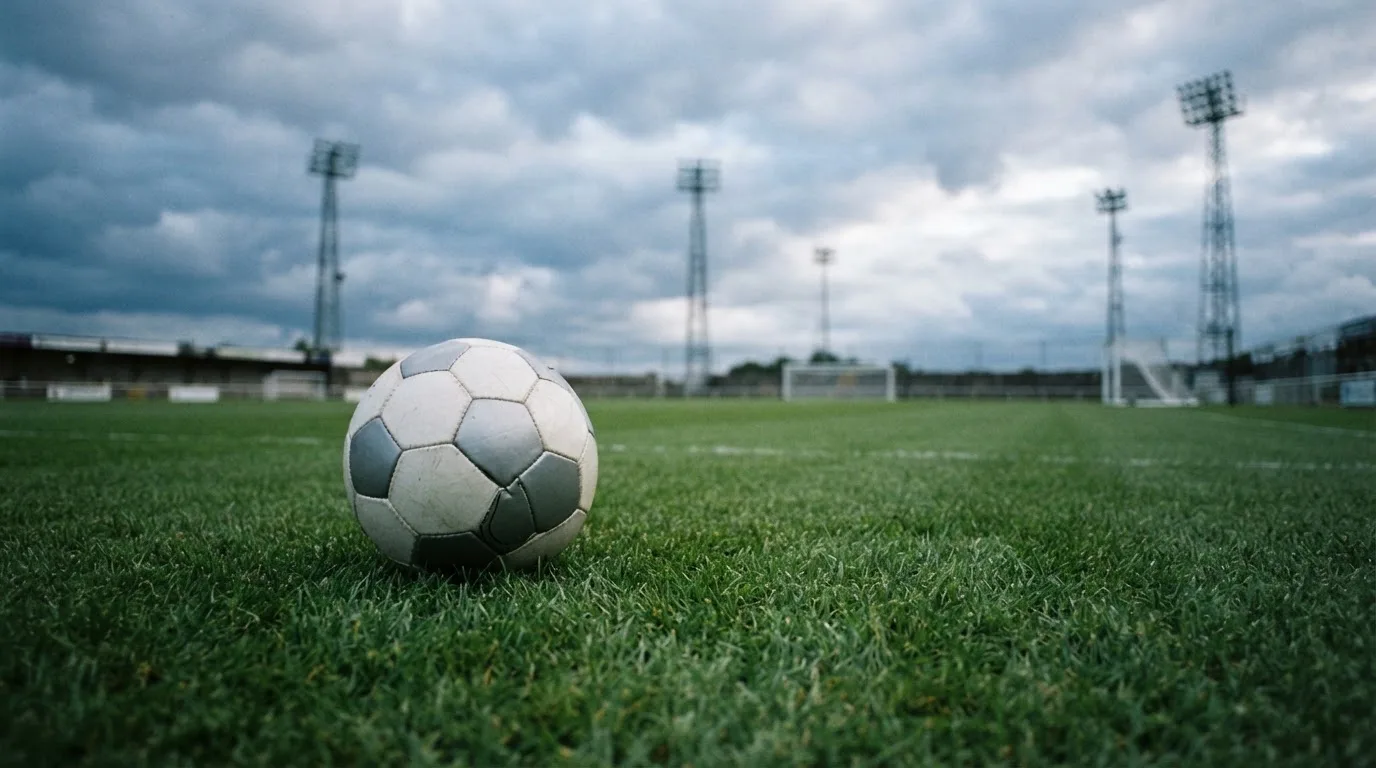Ein Fußball auf einem Rasenplatz, im Hintergrund Flutlichtmasten und ein bewölkter Himmel.