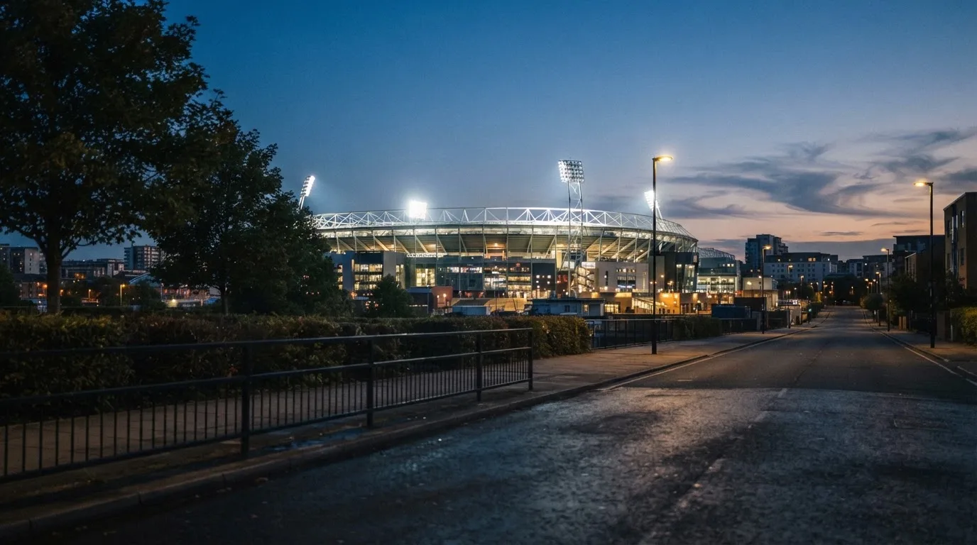Ein Stadion aus der Ferne bei Flutlicht, davor eine ruhige Straße in der Abenddämmerung.
