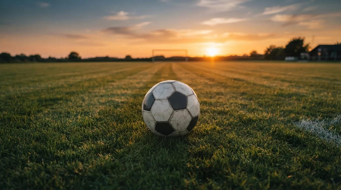 Ein Fußball liegt in der Mitte eines leeren Rasenplatzes, am Horizont färbt sich der Himmel orange.