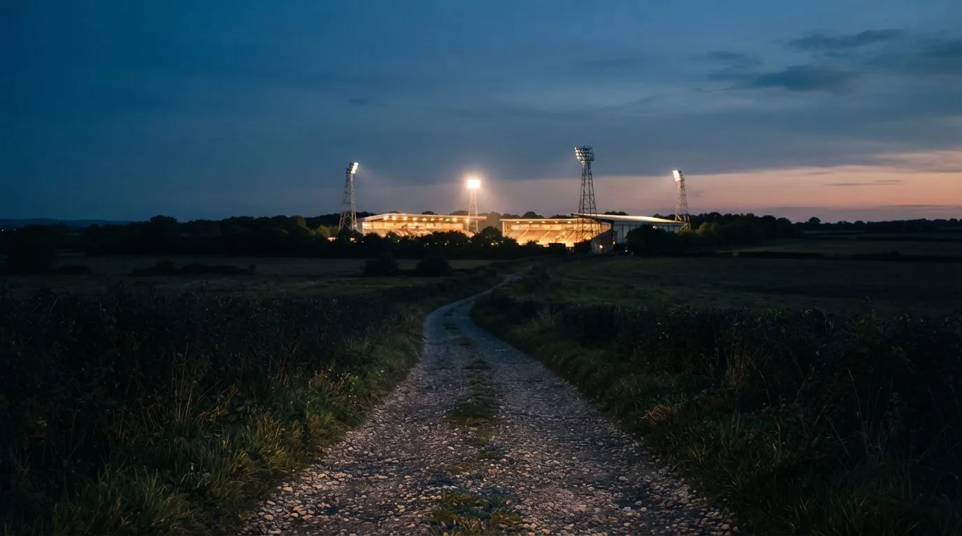 Ein beleuchtetes Stadion aus großer Distanz am Abend, im Vordergrund ein Feldweg.