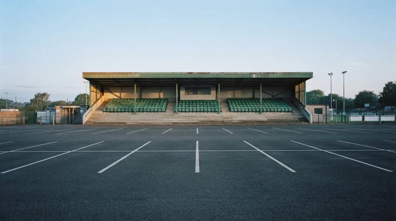 Eine leere Tribüne eines kleinen Stadions am frühen Morgen, aus der Ferne über einen menschenleeren Parkplatz fotografiert.