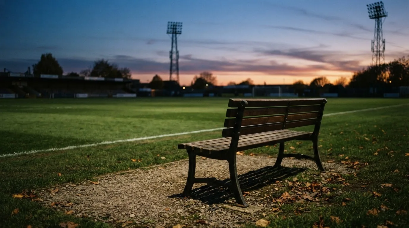 Eine leere Parkbank am Rand eines Fußballfelds in der Abenddämmerung, im Hintergrund ragen dunkle Flutlichtmasten in den Himmel.