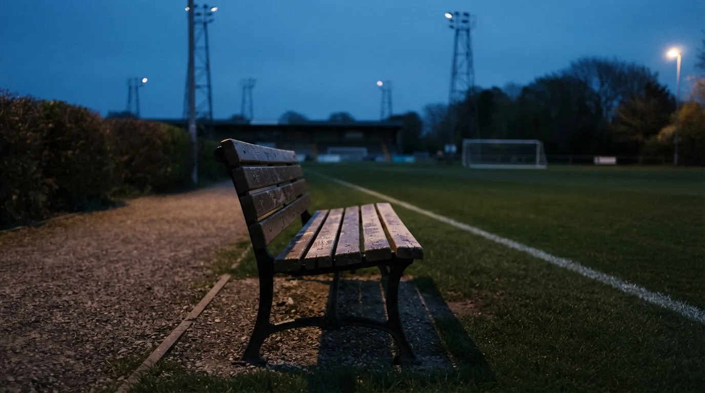 Eine leere Parkbank am Rand eines Fußballfelds in der Abenddämmerung, im Hintergrund ragen dunkle Flutlichtmasten in den Himmel.