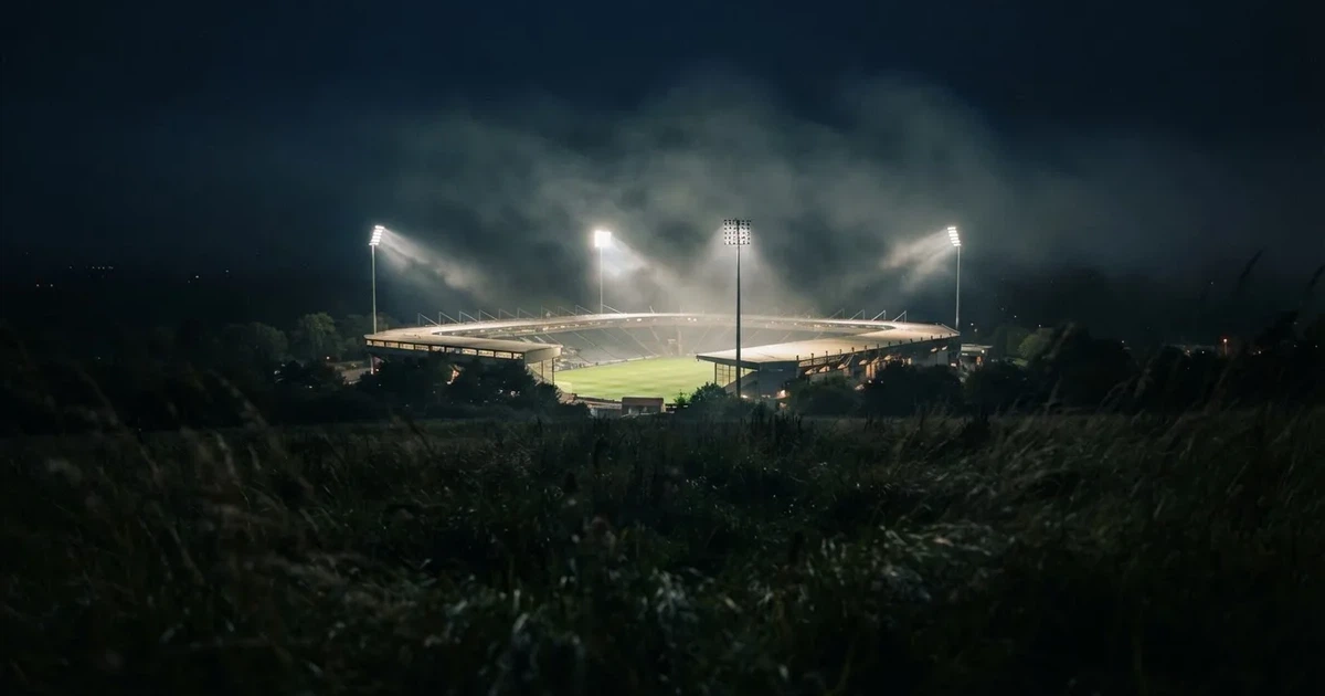 Ein Stadion unter Flutlicht, aus großer Entfernung über eine dunkle Wiese fotografiert, am Himmel zieht Nebel auf.