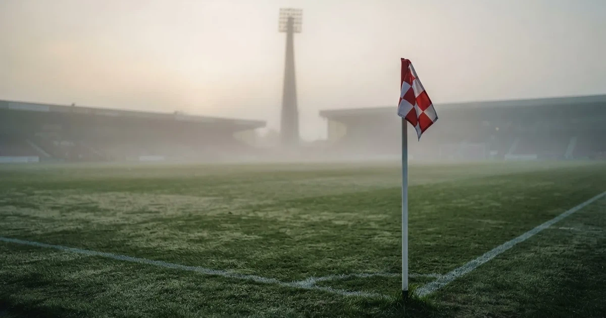 Eine Eckfahne steht im Nebel am Rand eines leeren Rasenplatzes in der Morgendämmerung, dahinter ein Flutlichtmast.