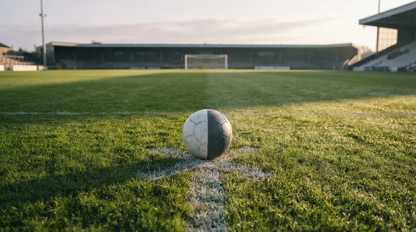 Ein Fußball liegt auf dem Anstoßpunkt eines leeren Rasenplatzes in der Morgendämmerung, im Hintergrund eine leere Tribüne.