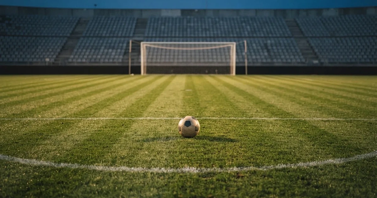Ein Fußballplatz mit frisch gemähtem Rasen im Abendlicht, ein Ball liegt im Strafraum, im Hintergrund unscharf eine leere Tribüne.