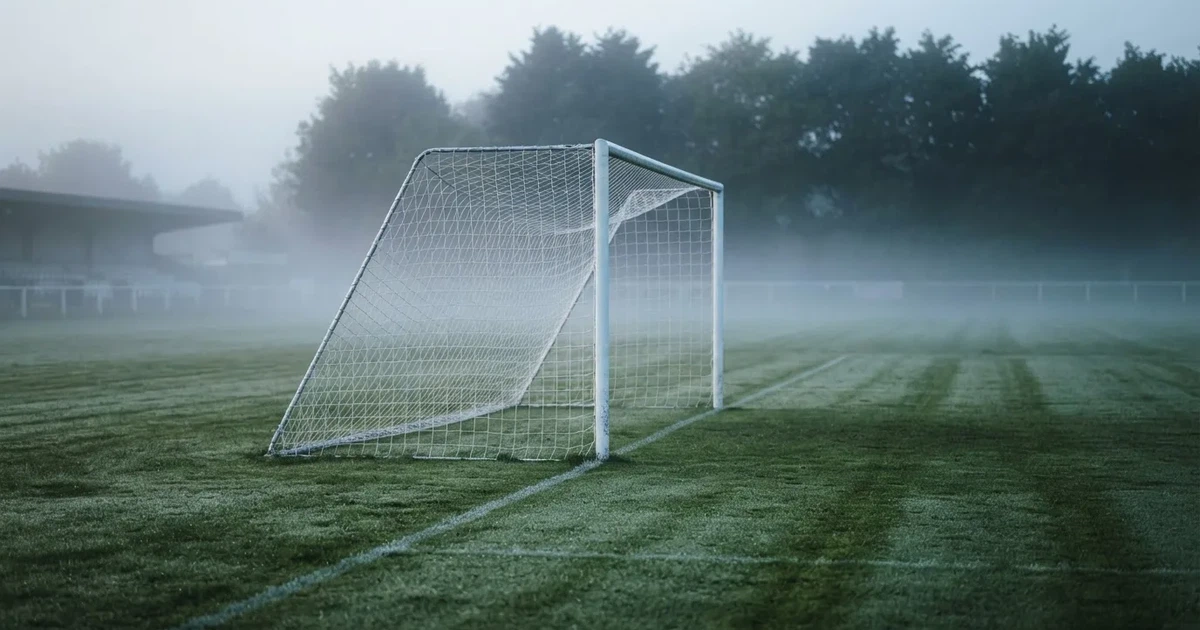 Ein Fußballtor mit Netz steht auf einem leeren Platz im Nebel der Morgendämmerung, der Rasen ist taufeucht.