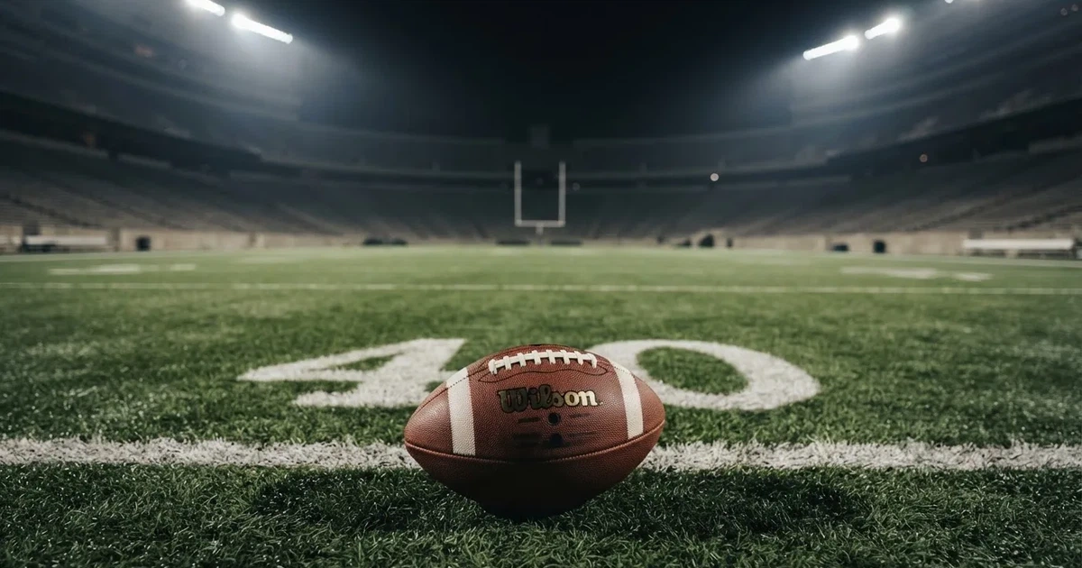 Ein American Football liegt auf dem Rasen eines leeren Stadions unter Flutlicht, die Torstangen ragen im Hintergrund auf.