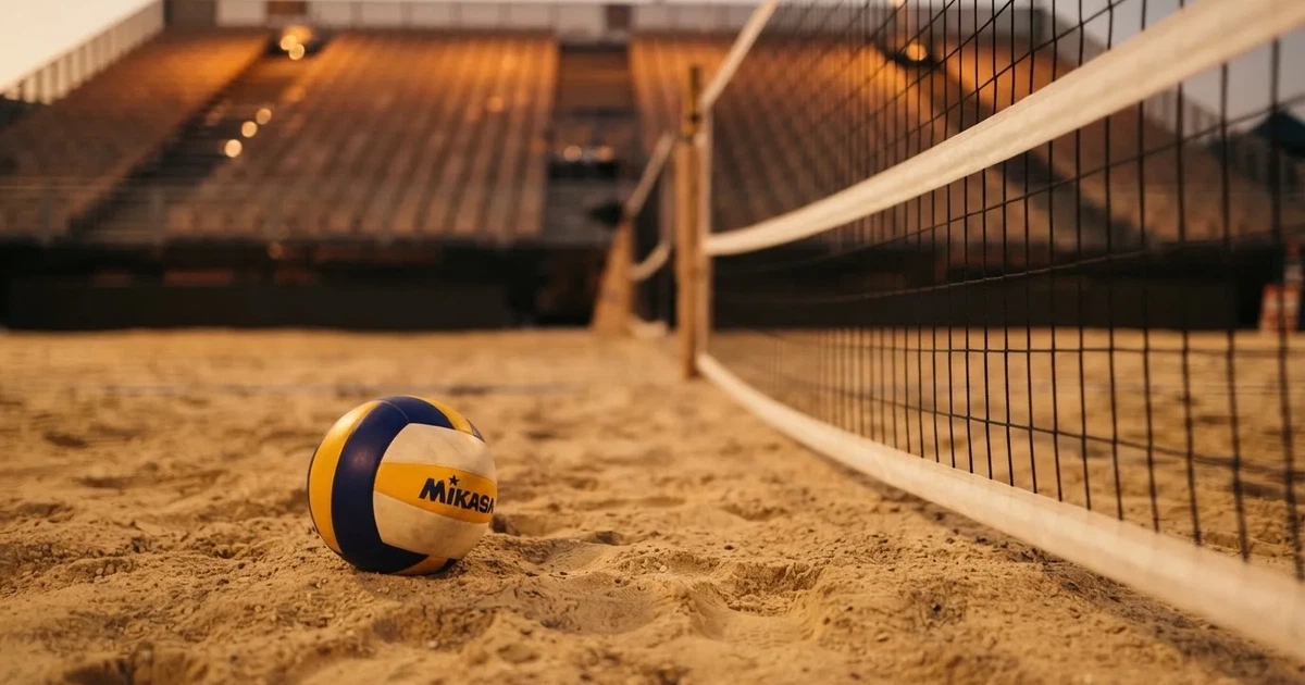 Ein Volleyball liegt auf dem Sand eines Beachvolleyball-Feldes neben dem gespannten Netz, im Hintergrund eine leere Tribüne bei Abendlicht.