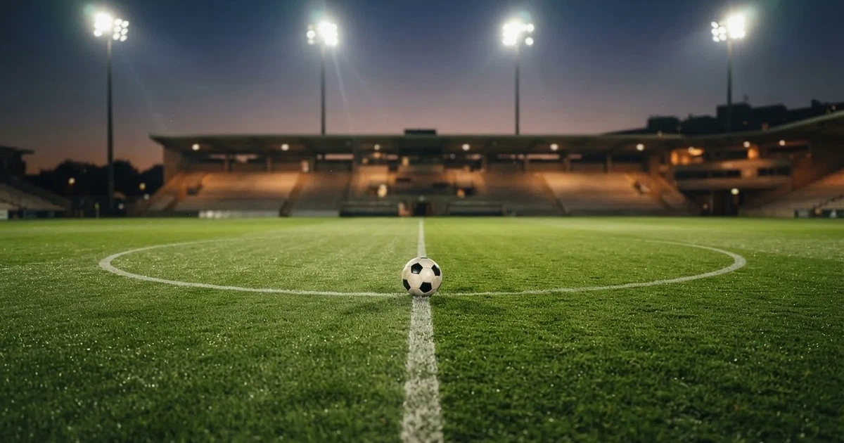 Ein Rasenplatz in der Dämmerung mit eingeschalteten Flutlichtmasten, ein Ball liegt am Anstoßpunkt, die Tribünen im Hintergrund sind unscharf.