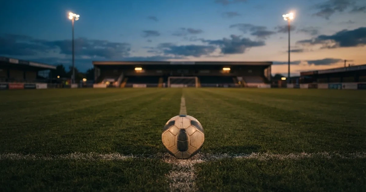 Ein Fußball liegt auf dem Anstoßpunkt eines leeren Rasenplatzes, im Hintergrund eine Tribüne und Flutlichtmasten in der Abenddämmerung.