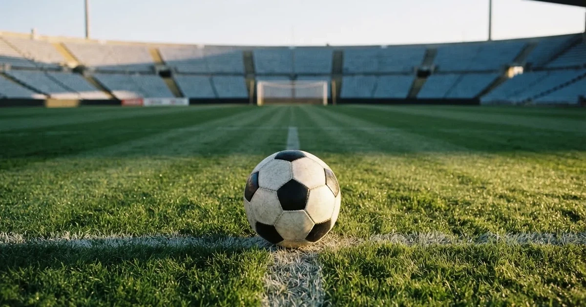 Ein Fußball liegt auf dem Anstoßpunkt eines Rasenplatzes, dahinter leere Tribünen im Licht des frühen Morgens.