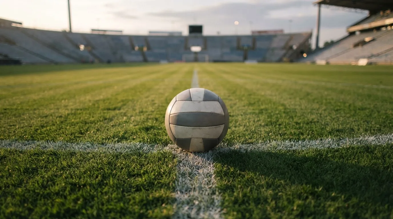 Ein Fußball liegt auf dem Anstoßpunkt eines Rasenplatzes, dahinter eine leere Tribüne im Abendlicht.