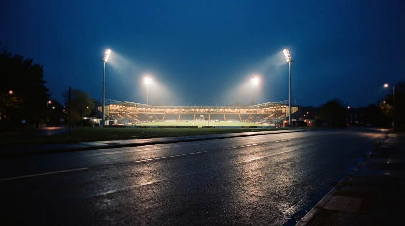 Ein Stadion unter Flutlicht aus großer Entfernung, davor eine leere Straße in der Abenddämmerung.