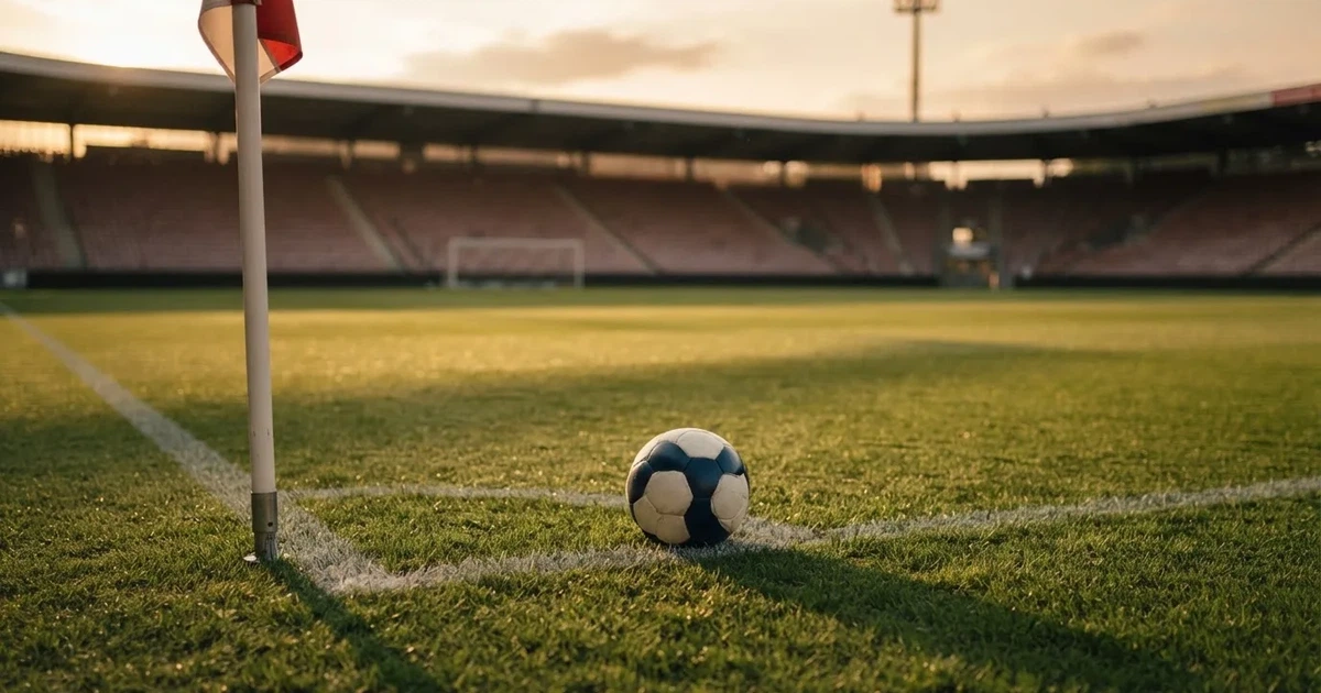 Ein Fußball liegt am Viertelkreis an der Eckfahne eines Rasenplatzes, im Hintergrund leere Tribünen in warmem Abendlicht.
