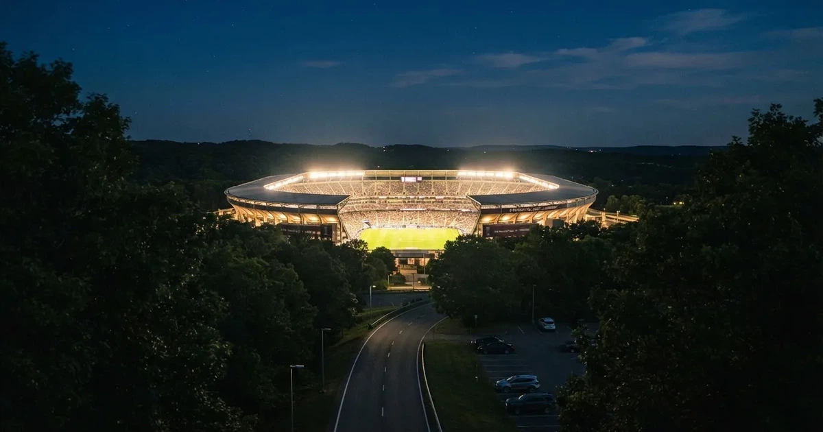 Ein Fußballstadion bei Nacht aus großer Entfernung, hell erleuchtet unter Flutlicht, davor eine dunkle Straße und Bäume.