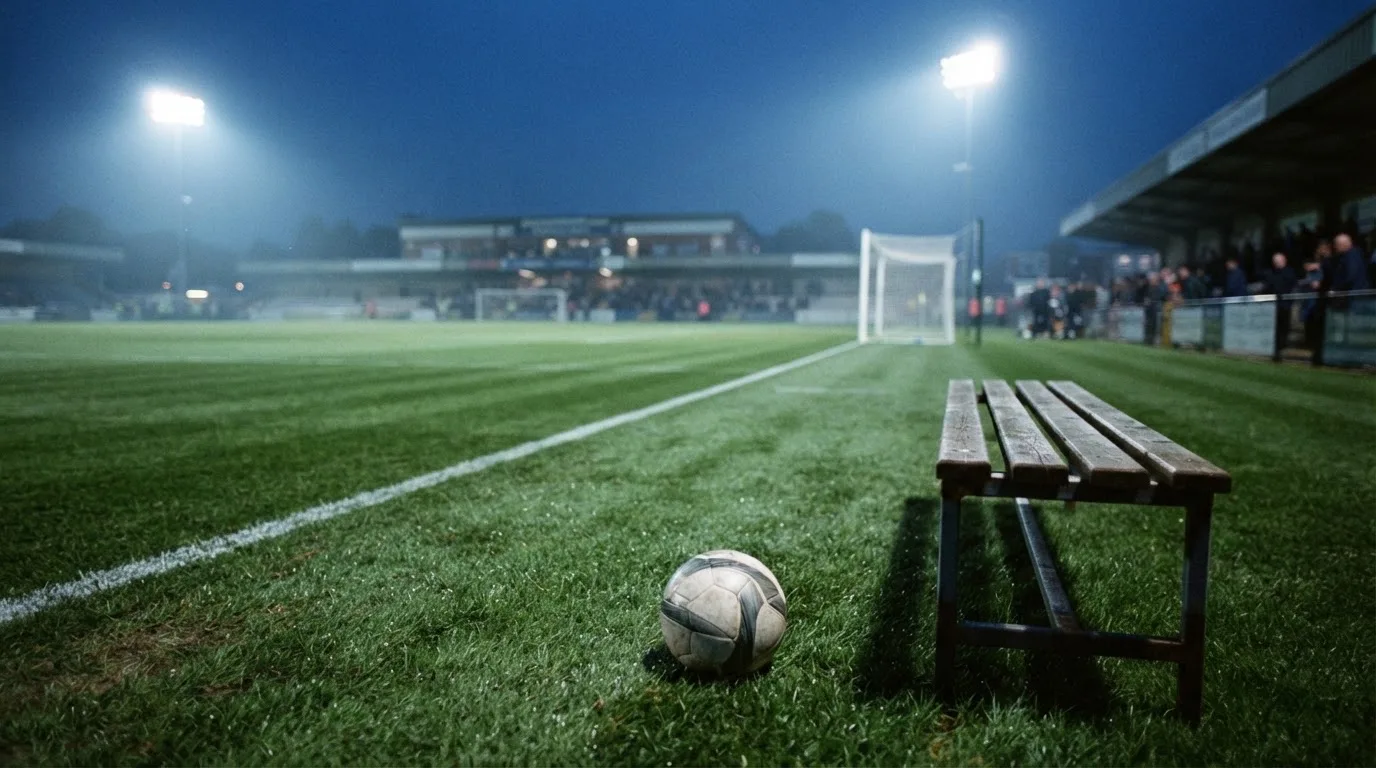 Ein Fußball liegt am Spielfeldrand neben einer leeren Ersatzbank, das Flutlicht über dem Platz leuchtet in der Dämmerung.
