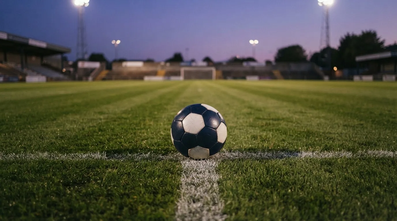 Ein Fußball liegt auf dem Anstoßpunkt eines Rasenplatzes in der Abenddämmerung, Flutlichtmasten stehen unscharf im Hintergrund.