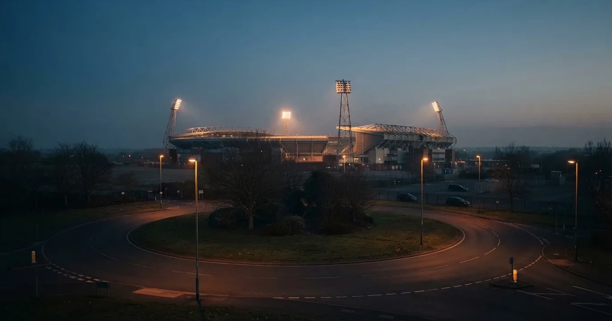 Ein Fußballstadion in der Dämmerung aus der Ferne, die Flutlichter gerade eingeschaltet, davor ein Kreisverkehr mit Laternen.