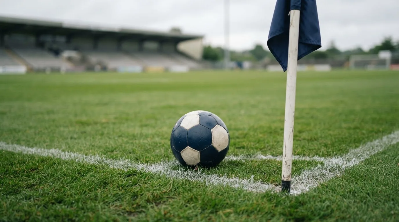 Ein Fußball liegt neben einer Eckfahne auf kurz gemähtem Rasen unter bewölktem Himmel.